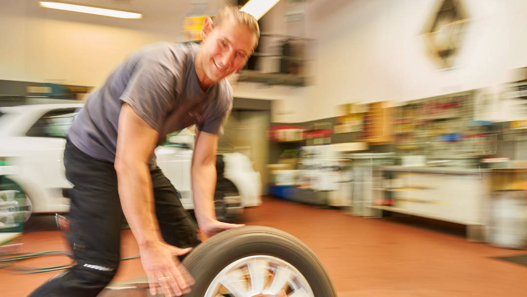 Ein lächelnder Mechaniker in einer Werkstatt rollt ein Autoreifenrad, während im Hintergrund unscharf ein weißes Auto und Werkstattregale zu sehen sind.