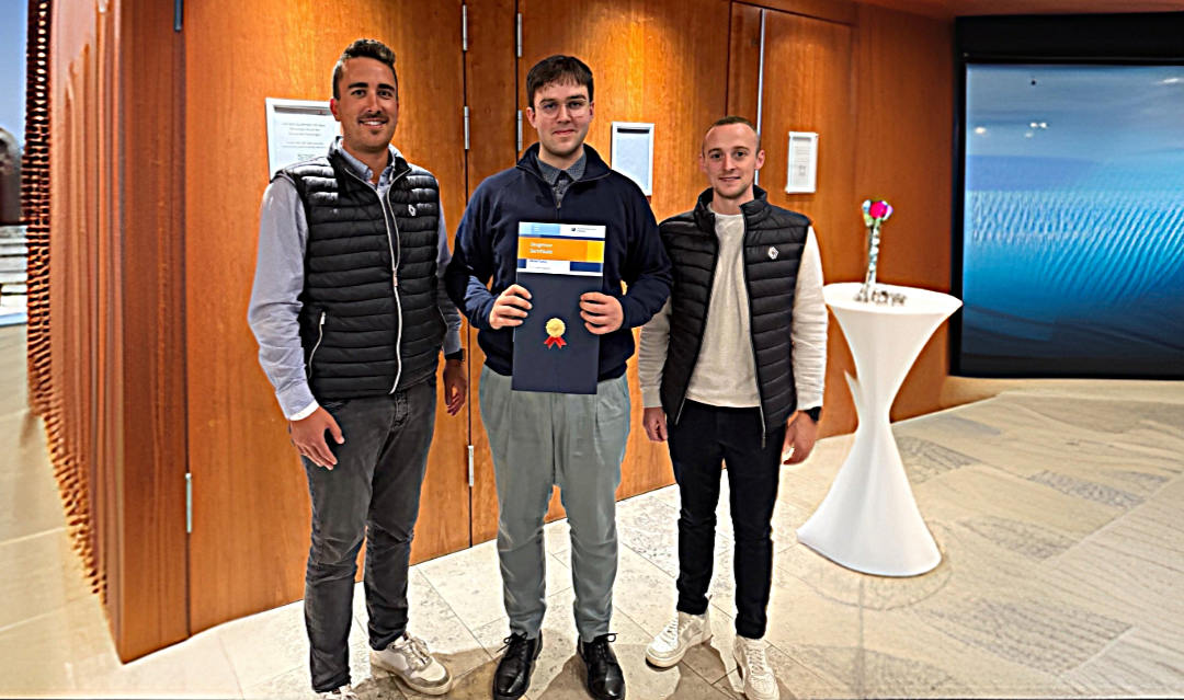 Luca Gutmann und Dennis Steck stehen in einem modernen Raum mit Holzverkleidung. Daniel Fuchs in der Mitte hält eine Urkunde in der Hand und lächelt. Alle drei tragen Freizeitkleidung, zwei von ihnen schwarze Steppwesten. Im Hintergrund steht ein Stehtisch mit einer Vase und einer Blume.