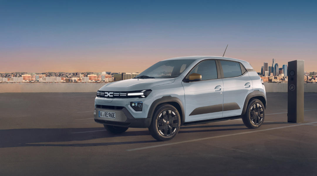 Ein kompakter, hellblauer Dacia Spring steht auf einem Parkdeck neben einer Ladesäule, im Hintergrund ist eine moderne Stadt-Skyline bei warmem Abendlicht zu sehen.