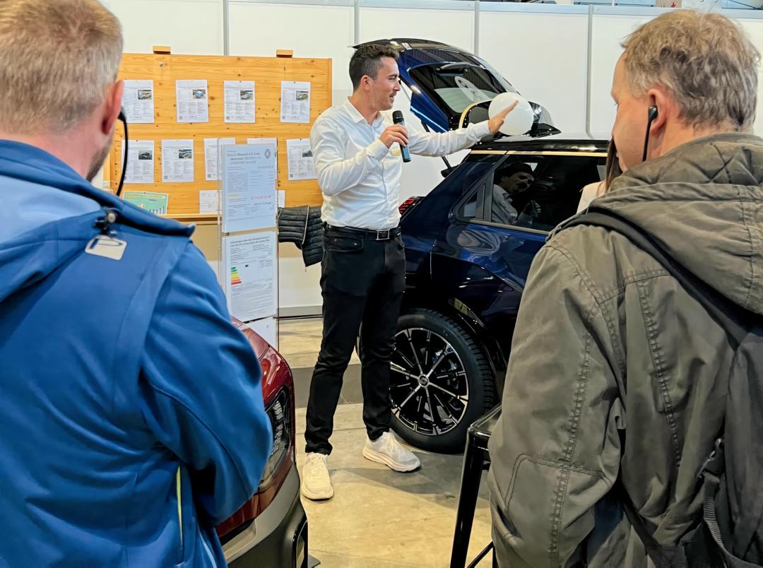 26-0301-getecIMG_4733 Luca Gutmann erklärt Besuchern ein ausgestelltes Fahrzeug mit geöffnetem Kofferraum während einer Präsentation auf der Messe.