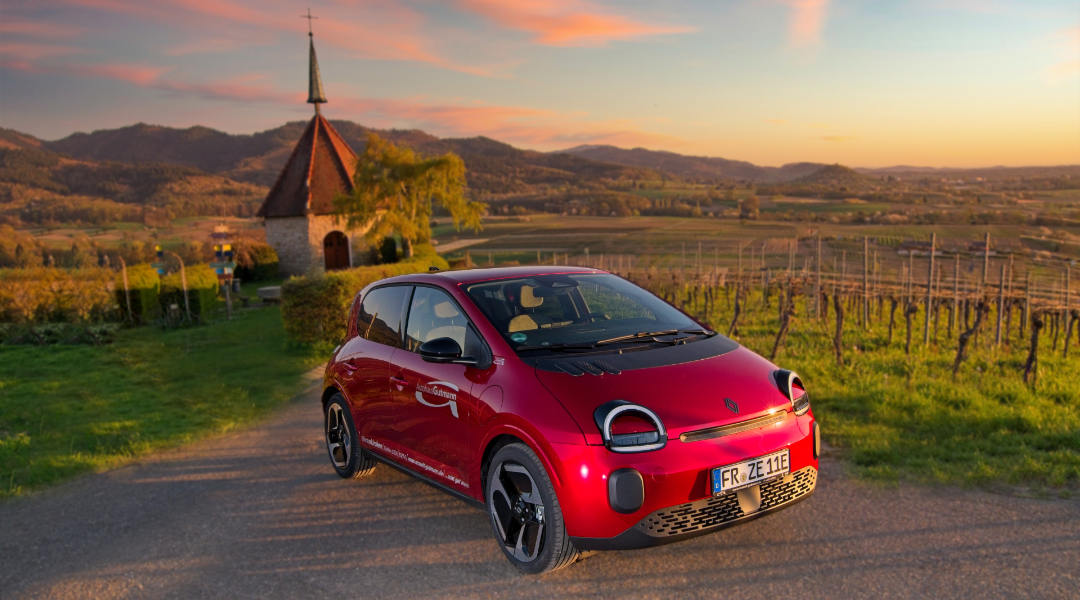 Ein roter Renault Twingo E-Tech, fährt auf einem schmalen Weg durch eine ländliche Weinberglandschaft bei Sonnenuntergang. Das Fahrzeug hat ein modernes Design mit runden Scheinwerfern und einem schwarzen Dach. Im Hintergrund sind Reihen von Weinreben zu sehen, die sich über sanfte Hügel erstrecken. Links im Bild befindet sich die Ölbergkapelle in Ehrenkirchen. Die Atmosphäre wirkt ruhig und idyllisch, mit warmen, goldenen Lichtstrahlen, die die Szene erhellen.