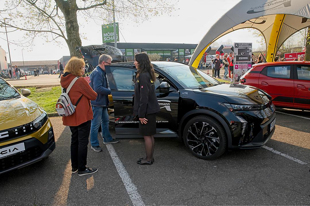Drei Personen stehen vor einem schwarzen Renault Scenic E-Tech mit geöffneten Türen am Stand des Autohauses Gutmann. Eine Verkäuferin in dunkler Jacke erklärt den beiden Kunden die Ausstattung des Fahrzeugs. Im Hintergrund sind weitere Autos zu sehen.