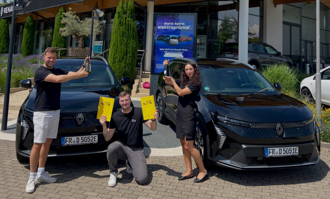 Drei lächelnde Personen posieren vor einem Autohaus zwischen zwei schwarzen Renault Scenic E-Tech Elektroautos. Sie halten feierlich Weinflaschen und zwei gelbe Übergabemappen in den Händen. Im Hintergrund ist ein Werbebildschirm mit der Aufschrift „liberté. égalité. elektroprämie*“ zu sehen.